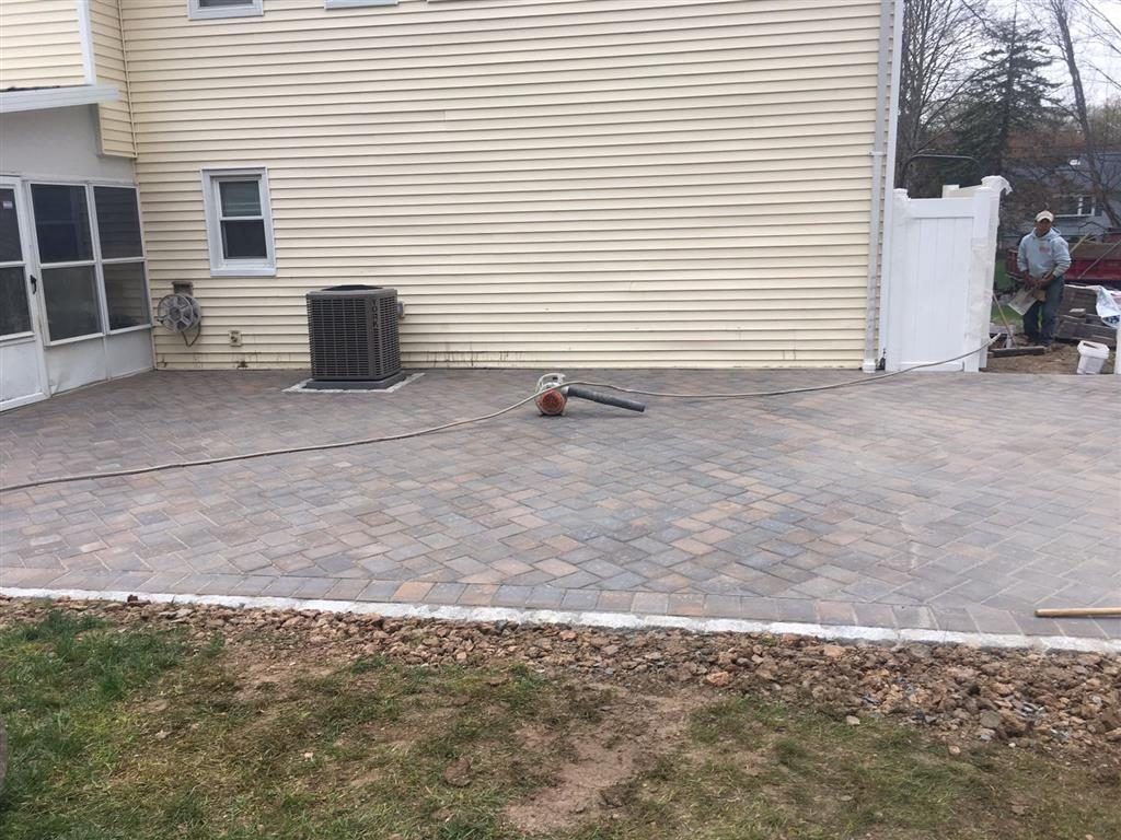 Brick-paved patio beside a beige house, with a leaf blower on the ground and lawn in front.