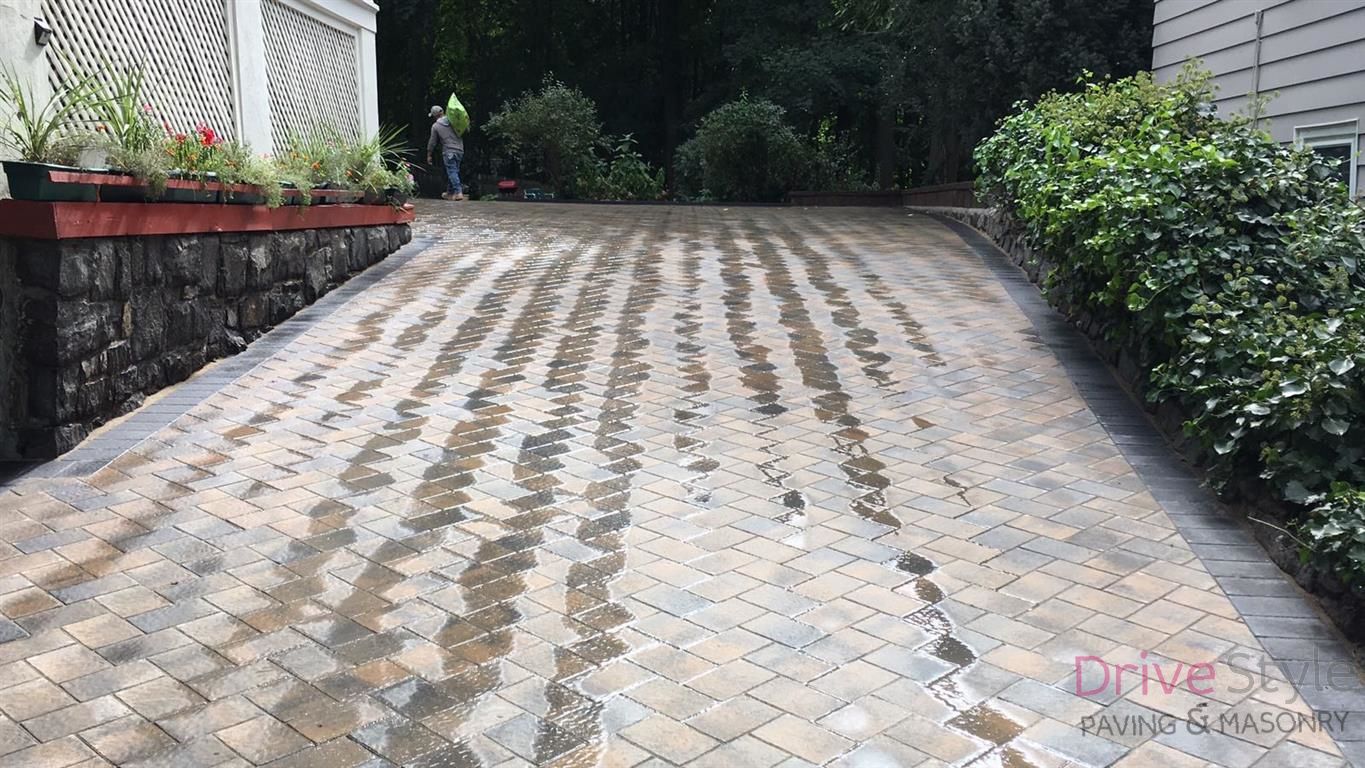 Paved driveway with interlocking bricks, bordered by greenery and a house wall on the left