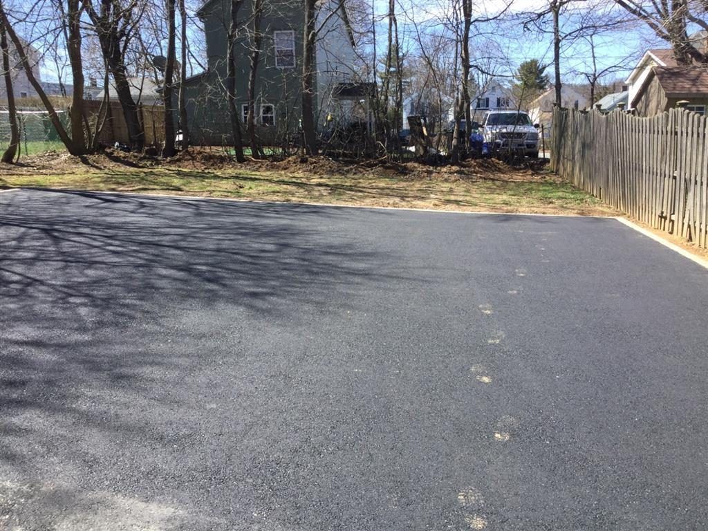 Paved driveway beside a wooden fence and trees, leading toward houses on a sunny day