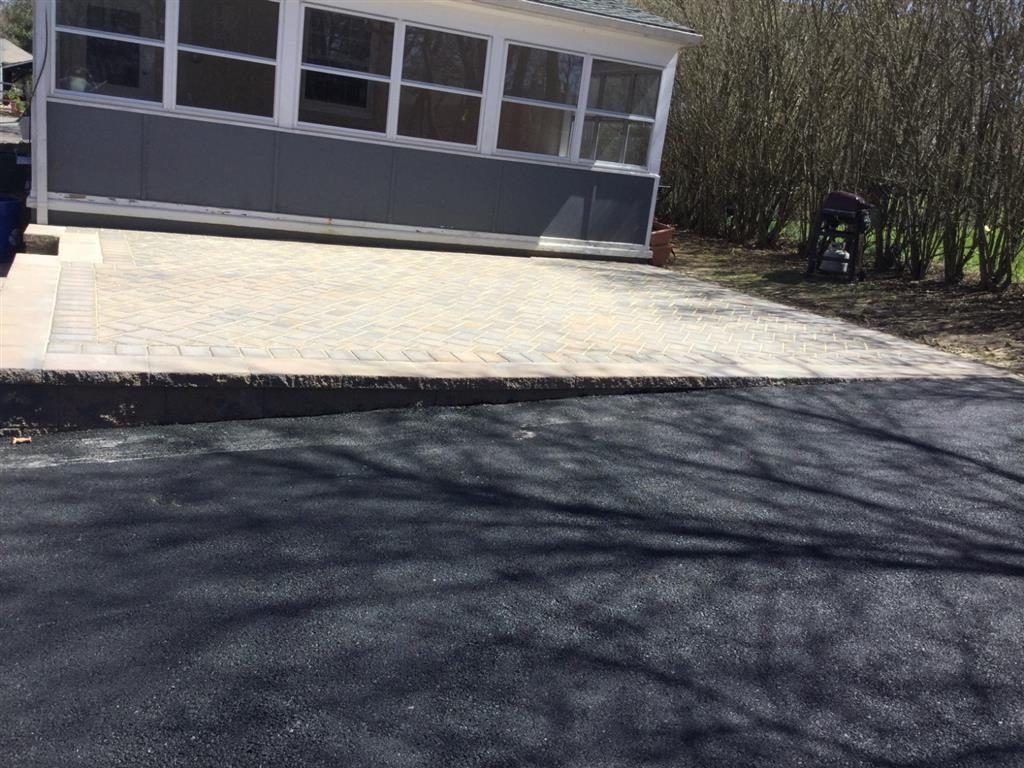 Sunlit driveway leading to a gray house with a screened porch and trees in the background