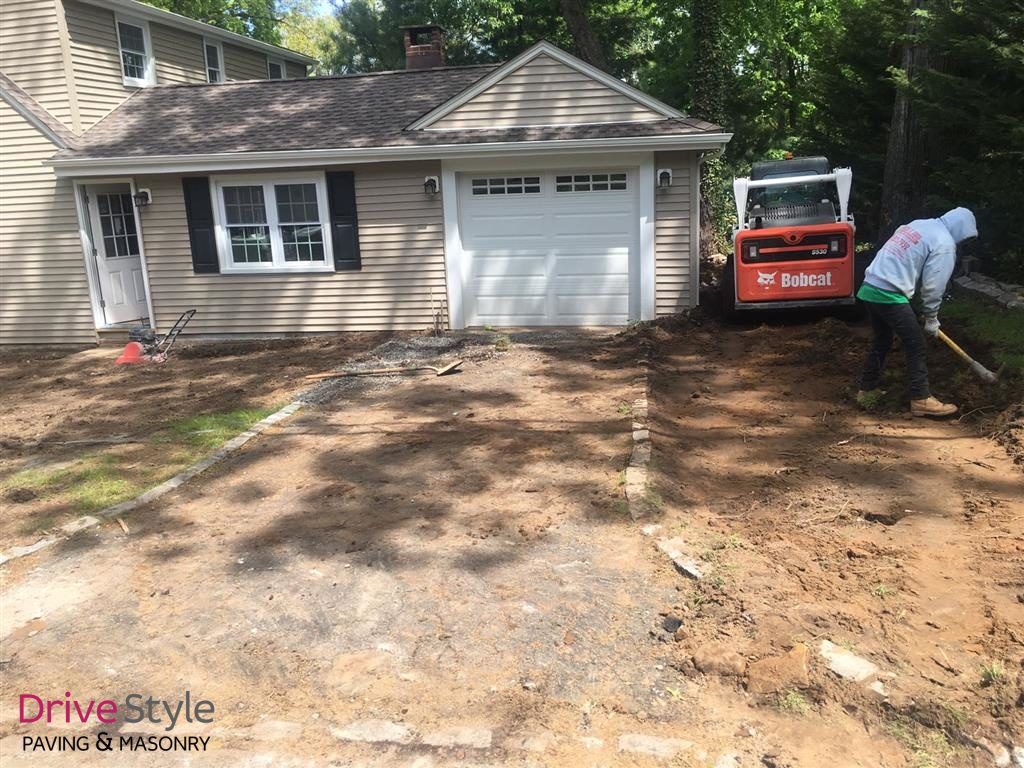 Driveway resurfacing beside a beige garage, with a worker using a shovel near a skid steer.