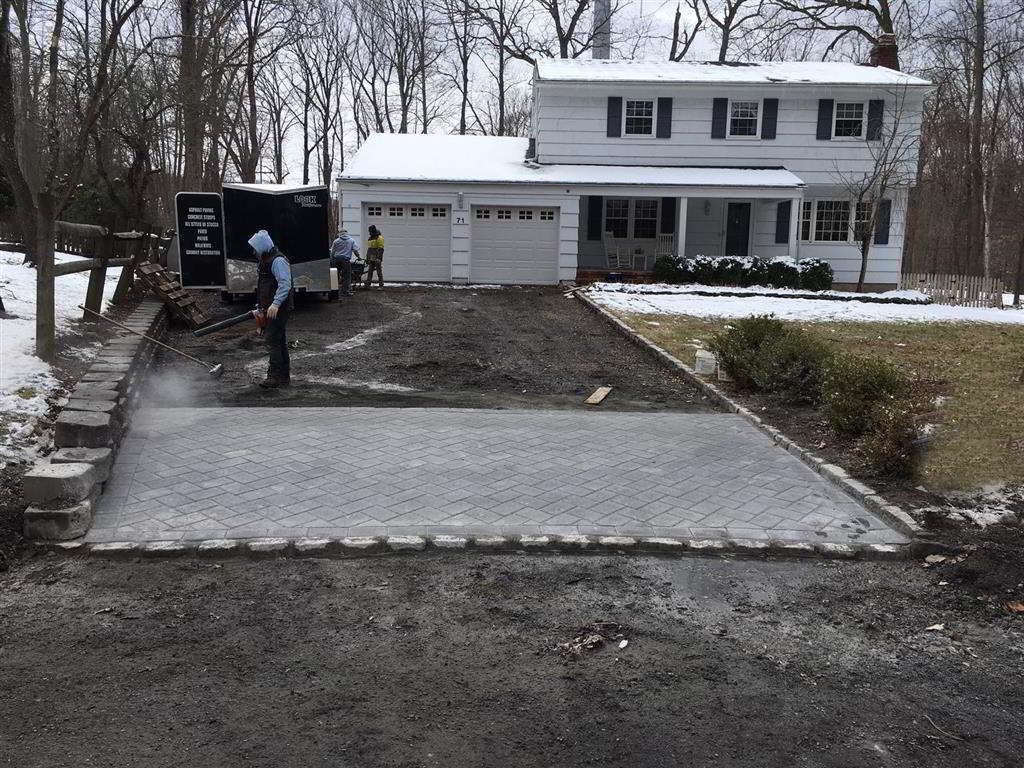 Driveway under construction leading to a white two-story house in a snowy yard, with one worker near a truck.
