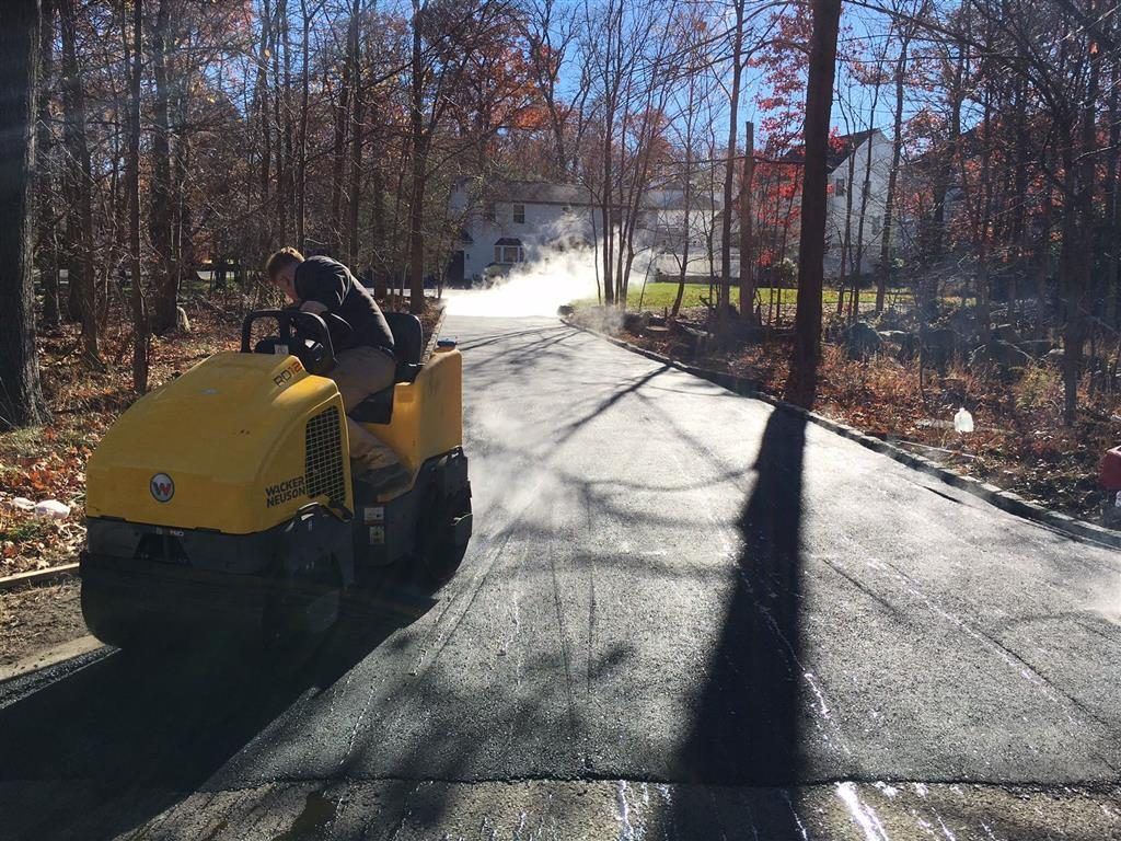 Yellow road roller paving a sunlit road in a wooded area