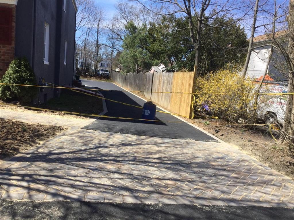 Sloped driveway beside a house and fence, with a child on a blue ride-on toy in the distance.