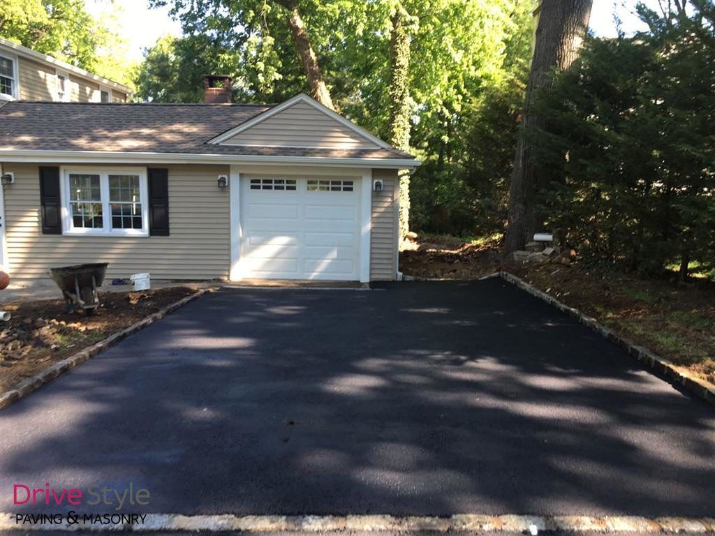 Freshly paved black driveway leading to a gray house with a white garage door and trees behind it
