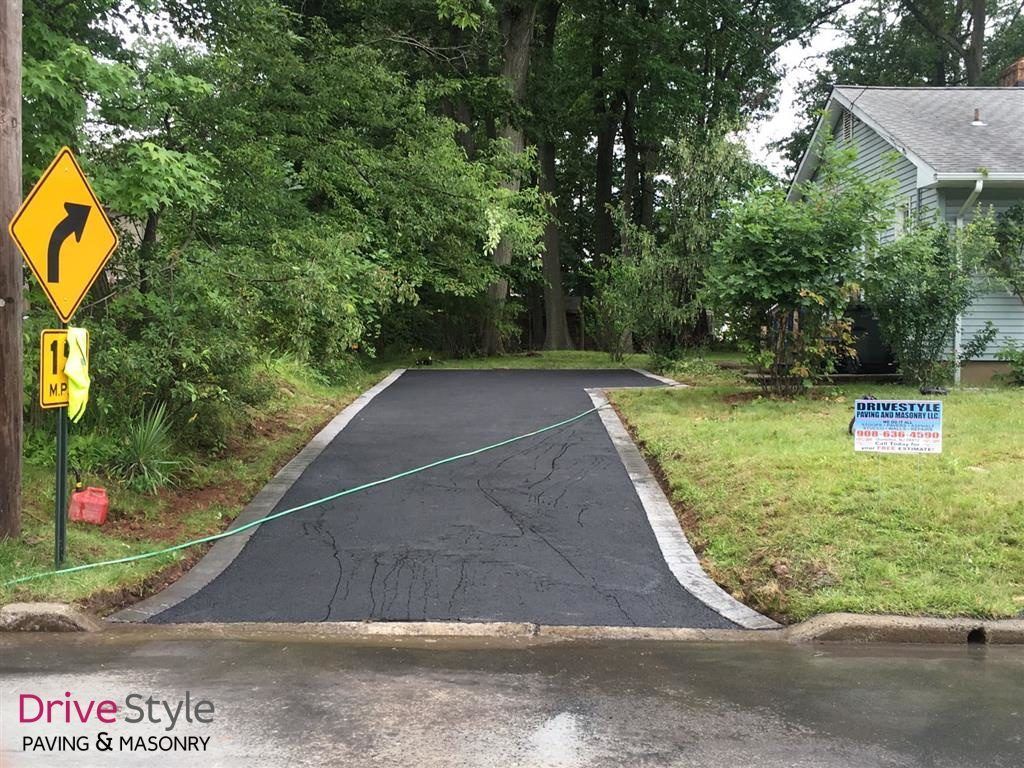 Driveway with cracked pavement and a blue caution tape across the entrance, surrounded by trees and a house.