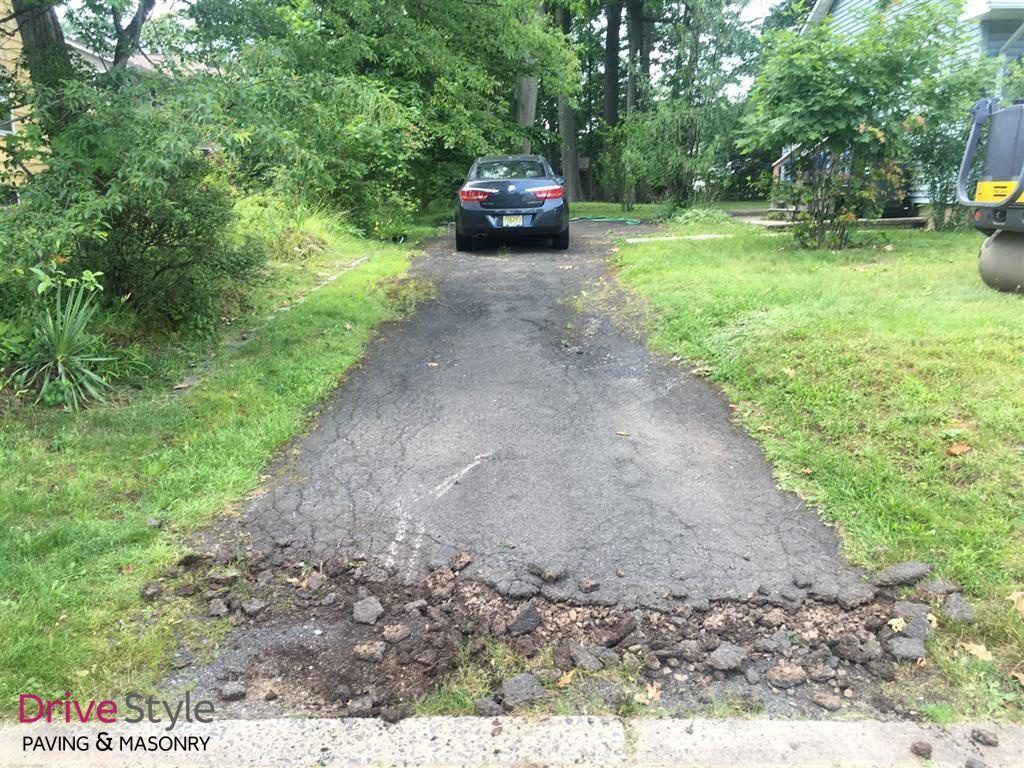 Black car on a narrow paved driveway with cracked asphalt, surrounded by green grass and trees.