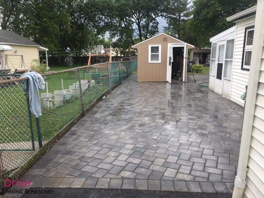 Paved backyard patio with a small shed, chain-link fence, and houses on both sides.