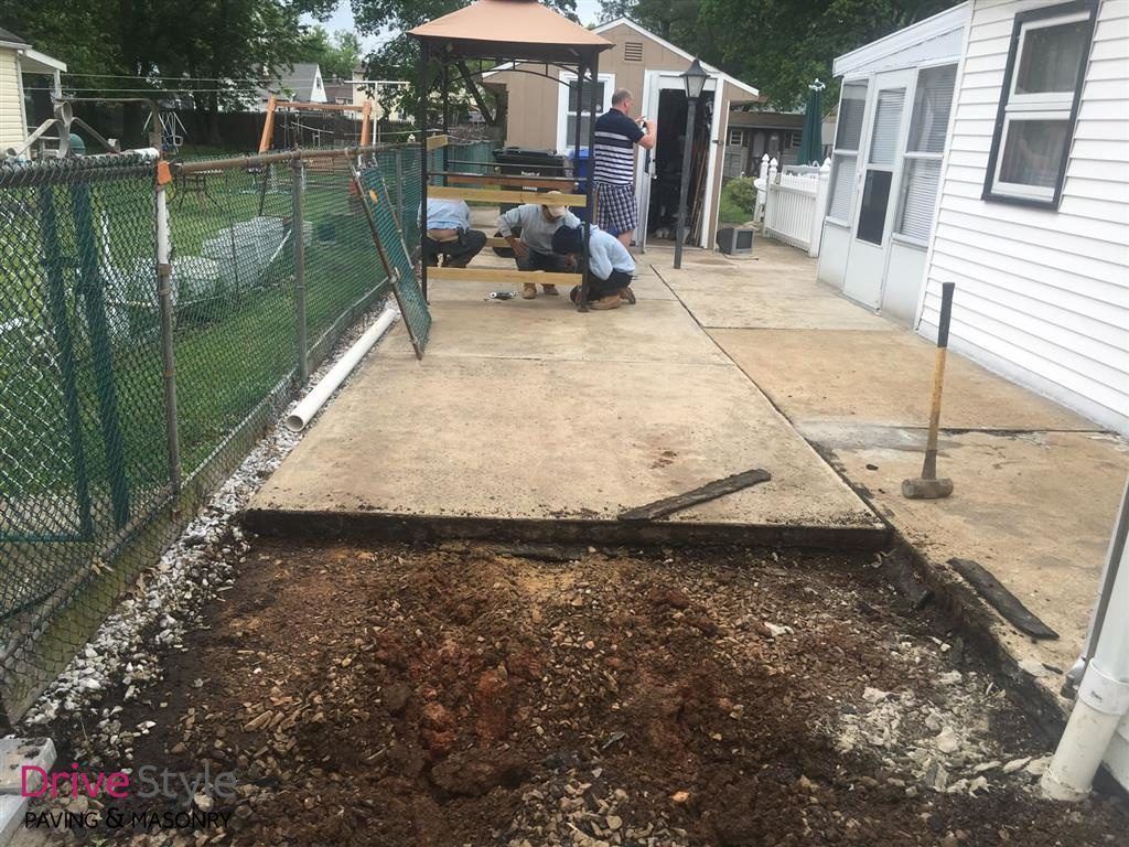 Two workers on a driveway beside a white house, removing a section of old pavement.