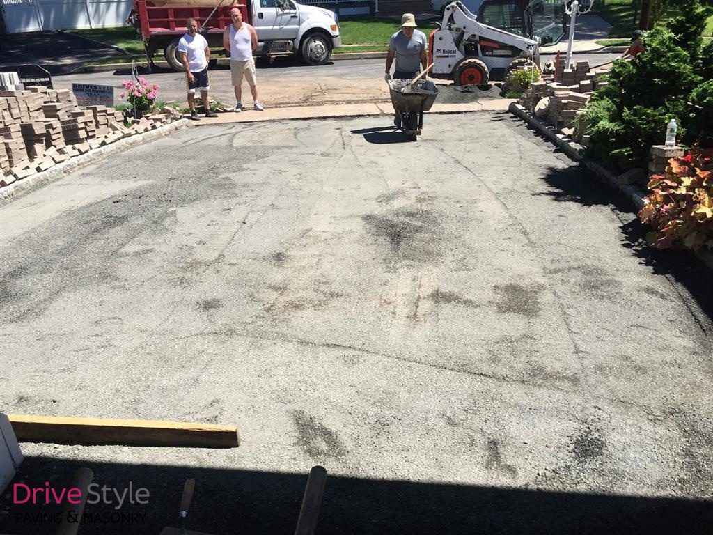 Workers paving a driveway, with one person pushing a wheelbarrow of asphalt in a sunny yard