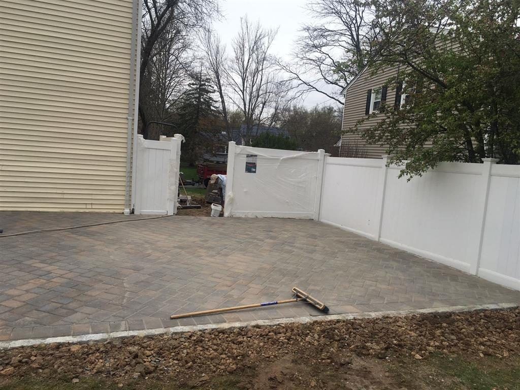 Paved backyard patio with white fence, beige house wall, and a fallen tool on the ground