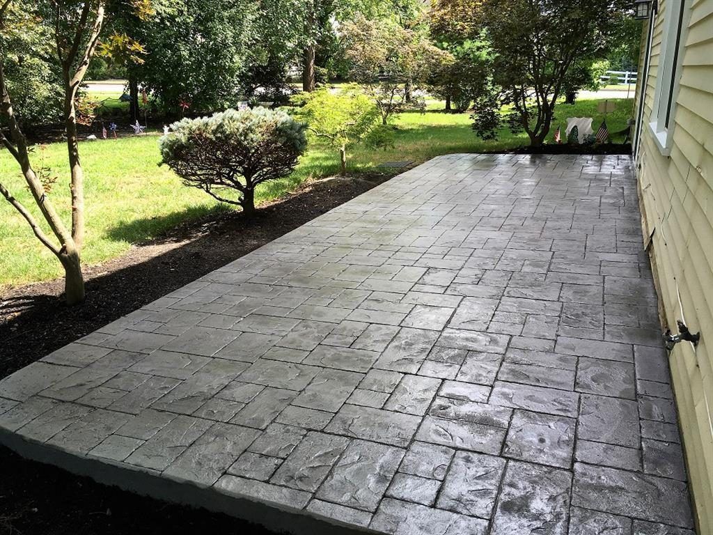 Paved patio beside a house, bordered by shrubs and trees in a sunny yard.
