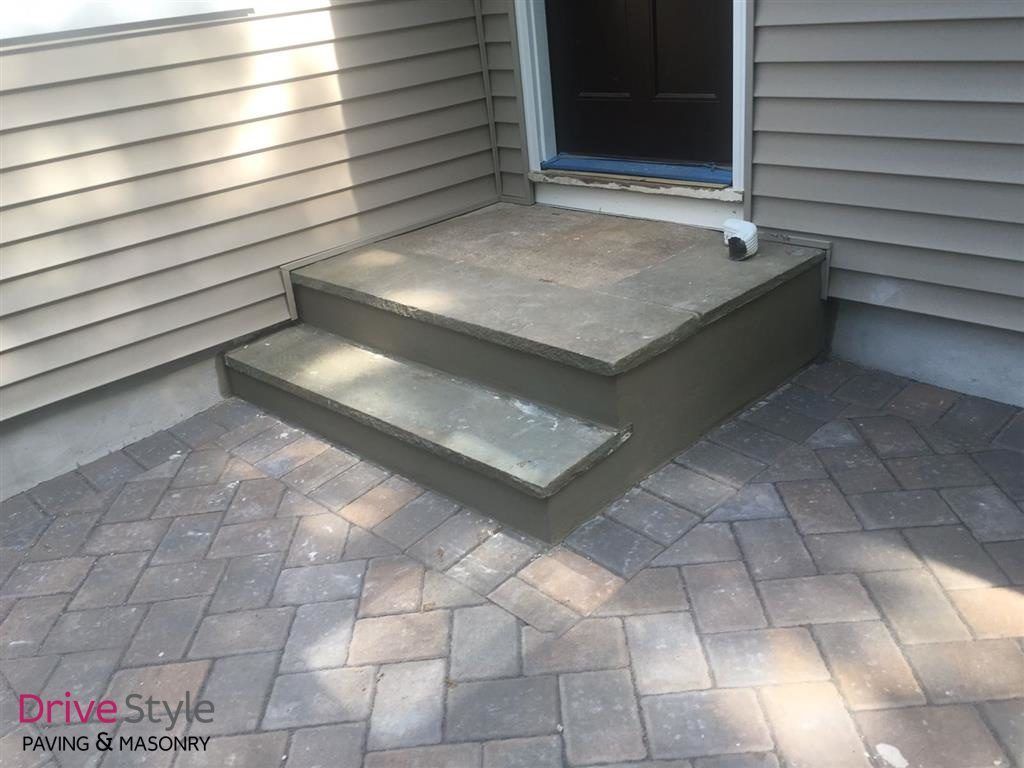 Small concrete porch with two steps leading to a doorway, beside gray siding and a brick-paver patio.