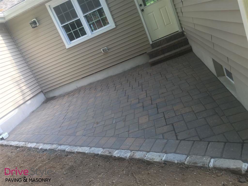 Paved brick patio beside beige house siding, with windows, door, and low curb edging.