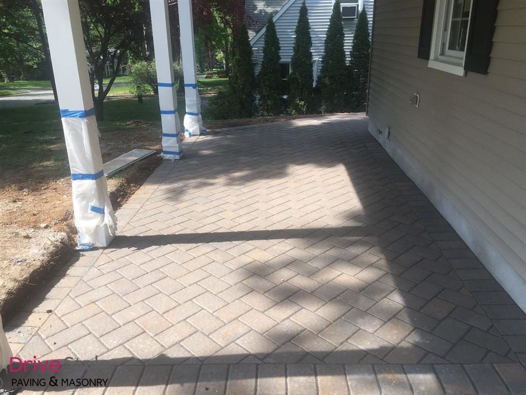 Paver patio beside a house with white columns, blue tape wrapped around them, and evergreen shrubs in back.