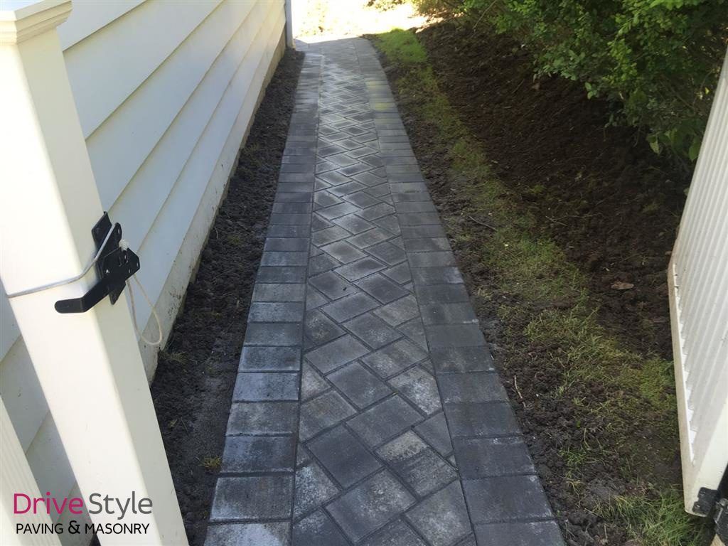 Narrow side walkway with gray pavers between a house and a hedge-lined fence.