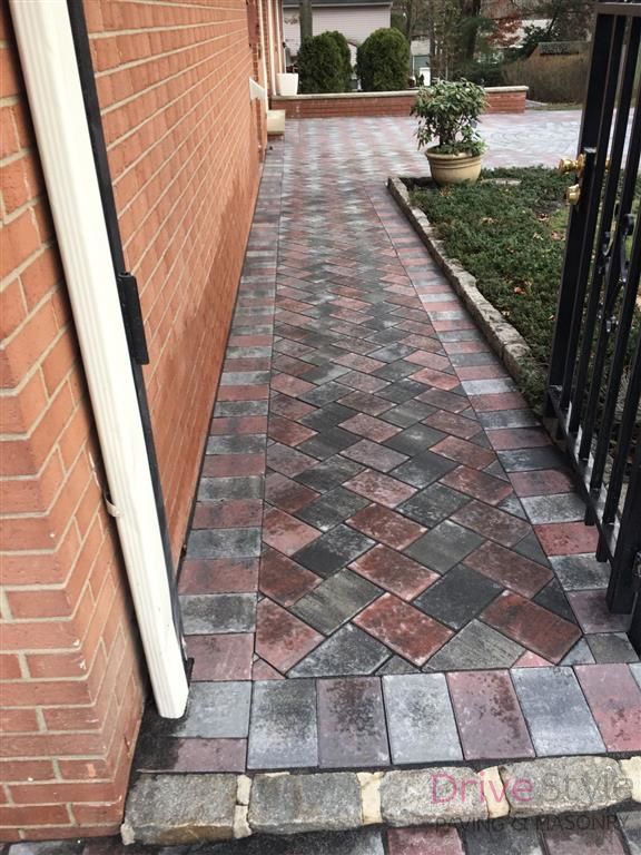 Brick walkway beside a house, paved with red and gray tiles and bordered by a black fence and shrubs.