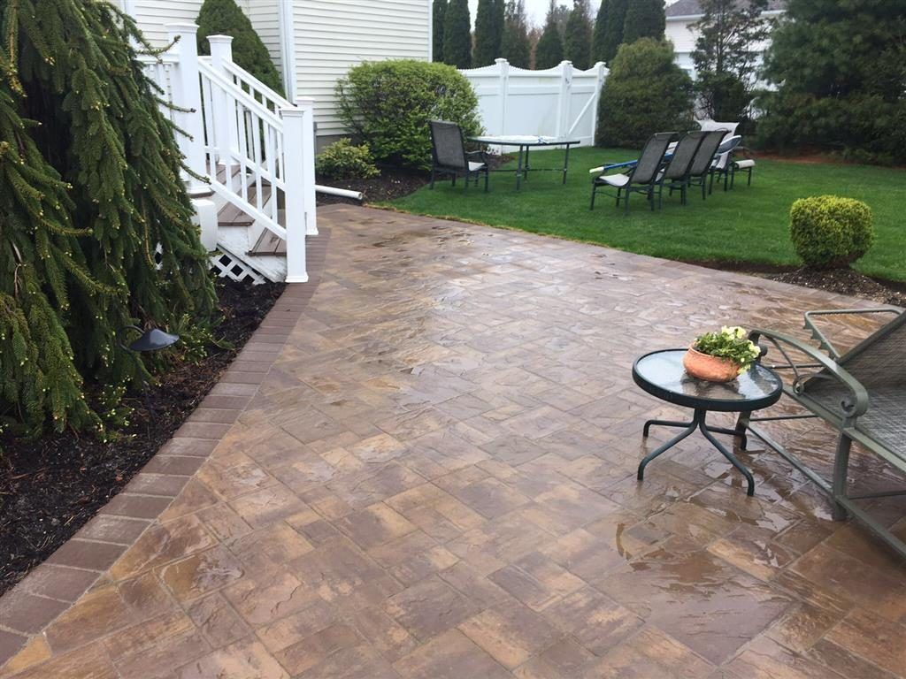 Wet brick patio beside white house, with outdoor chairs, table, and green lawn after rain.
