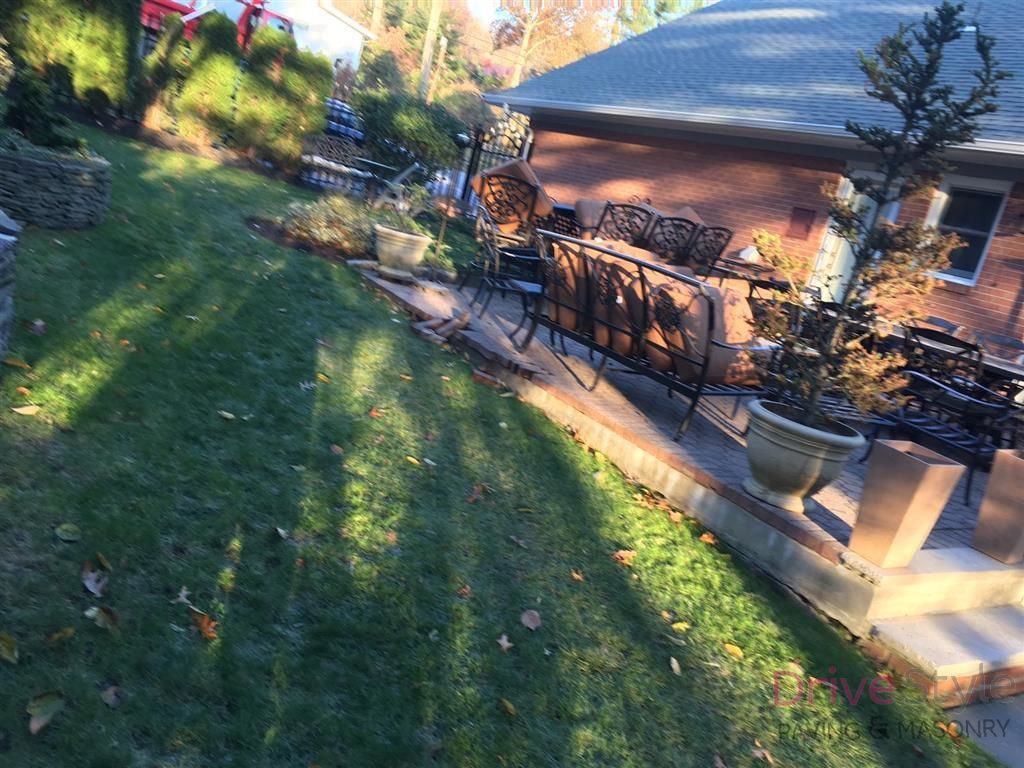 Sunlit backyard patio with black metal table and chairs beside a brick house, overlooking a grassy lawn