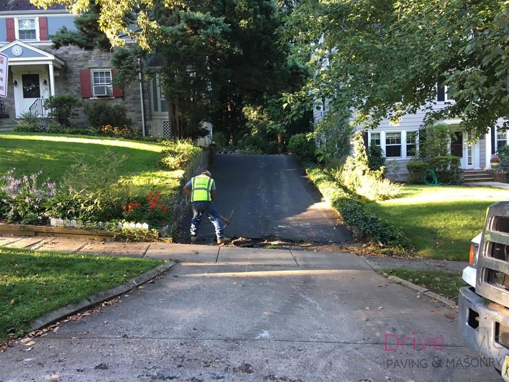 Worker paving a steep driveway between suburban houses, with a truck parked on the right.