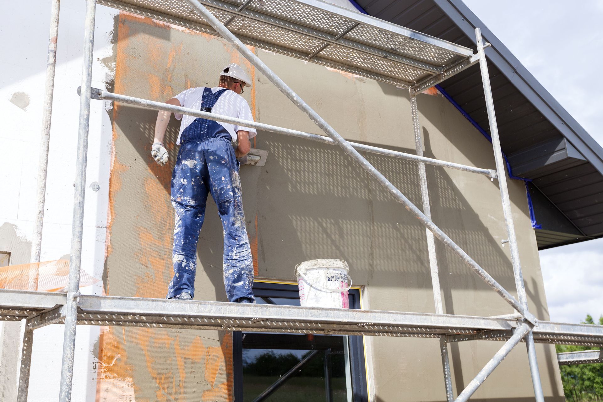 Painter on scaffolding applying orange coating to an exterior wall next to a window