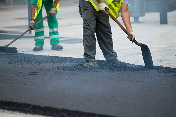 Workers paving a road, spreading hot asphalt with shovels and safety vests.