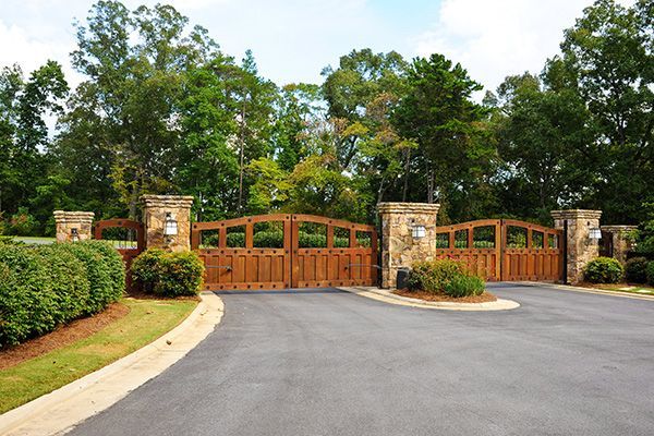Wooden gated entrance with stone pillars beside a curved paved driveway and green trees
