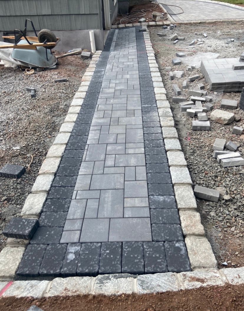 Narrow paver walkway under construction, with gray and black stones bordered by light edging.