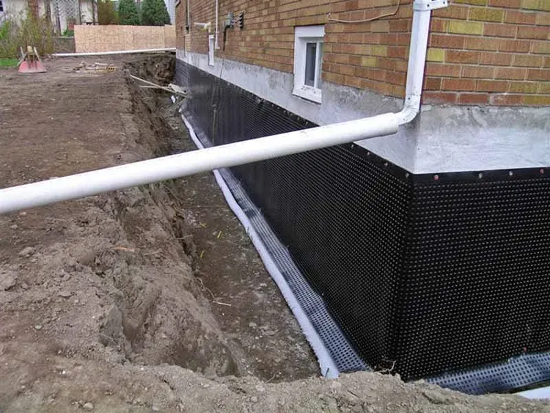 House foundation with black waterproofing, white downspout, and trench along the side of a brick home