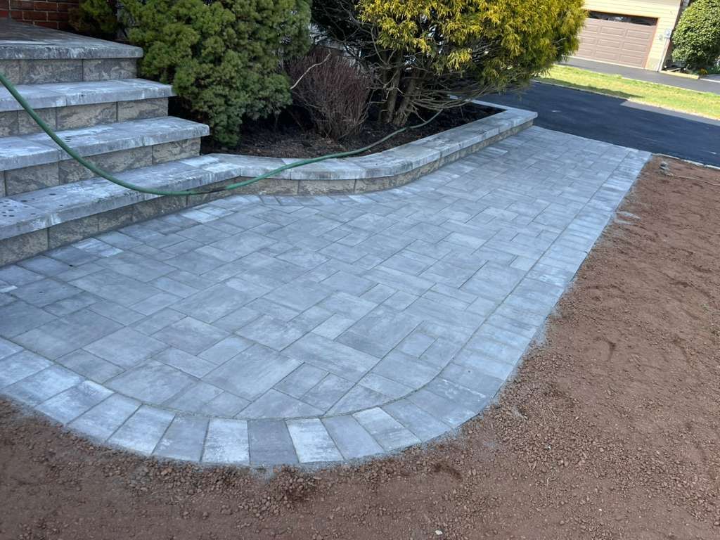 Curved stone paver walkway beside front steps and shrubs, bordered by soil and a street.