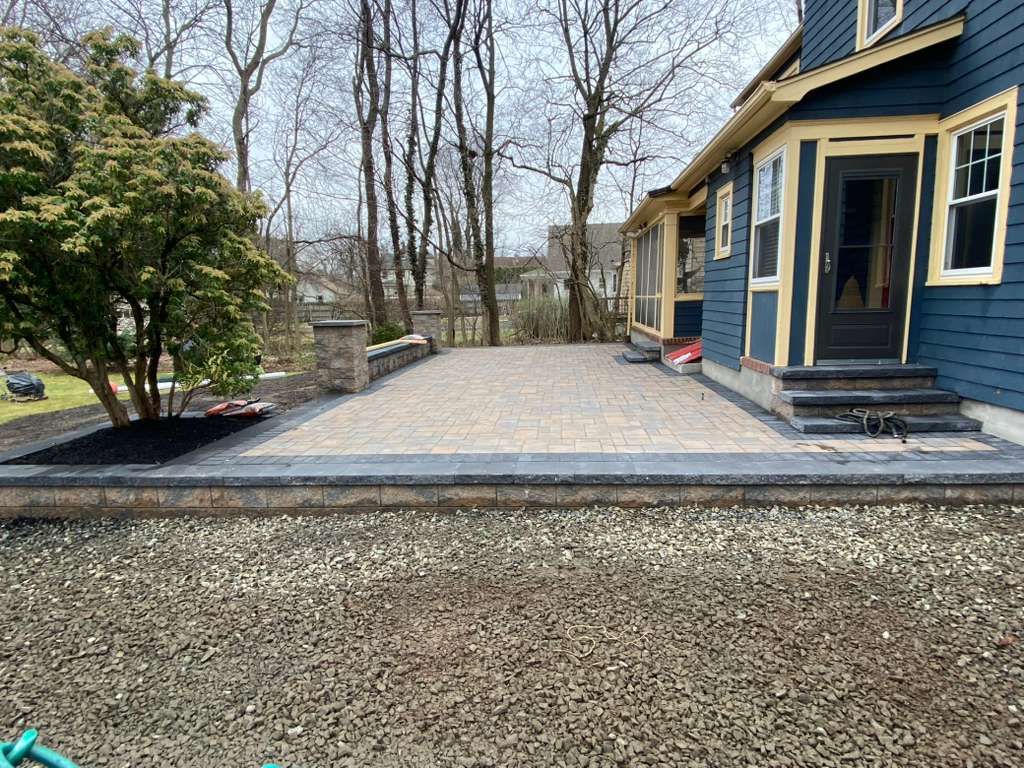 Blue house with a stone patio and gravel driveway, bordered by trees and shrubs.