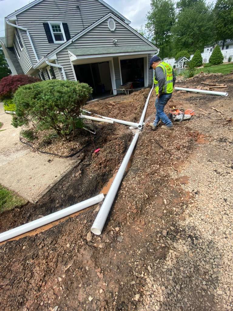 Worker in neon vest inspects broken white pipe near a house amid muddy yard damage.