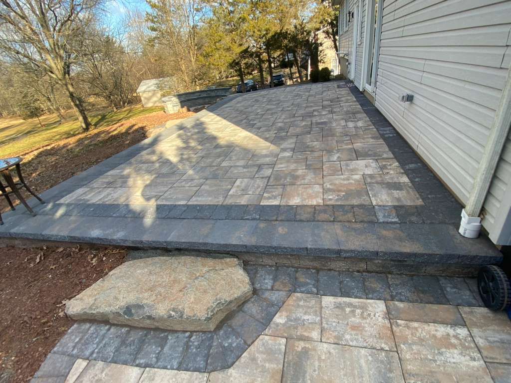 Paver patio with steps and a large stone, beside a white-sided house in a backyard.