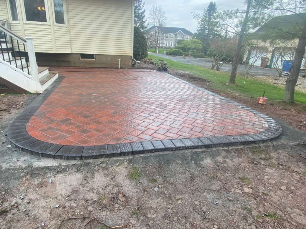 Red brick patio bordered by dark pavers beside a house, with wet ground in a residential yard.