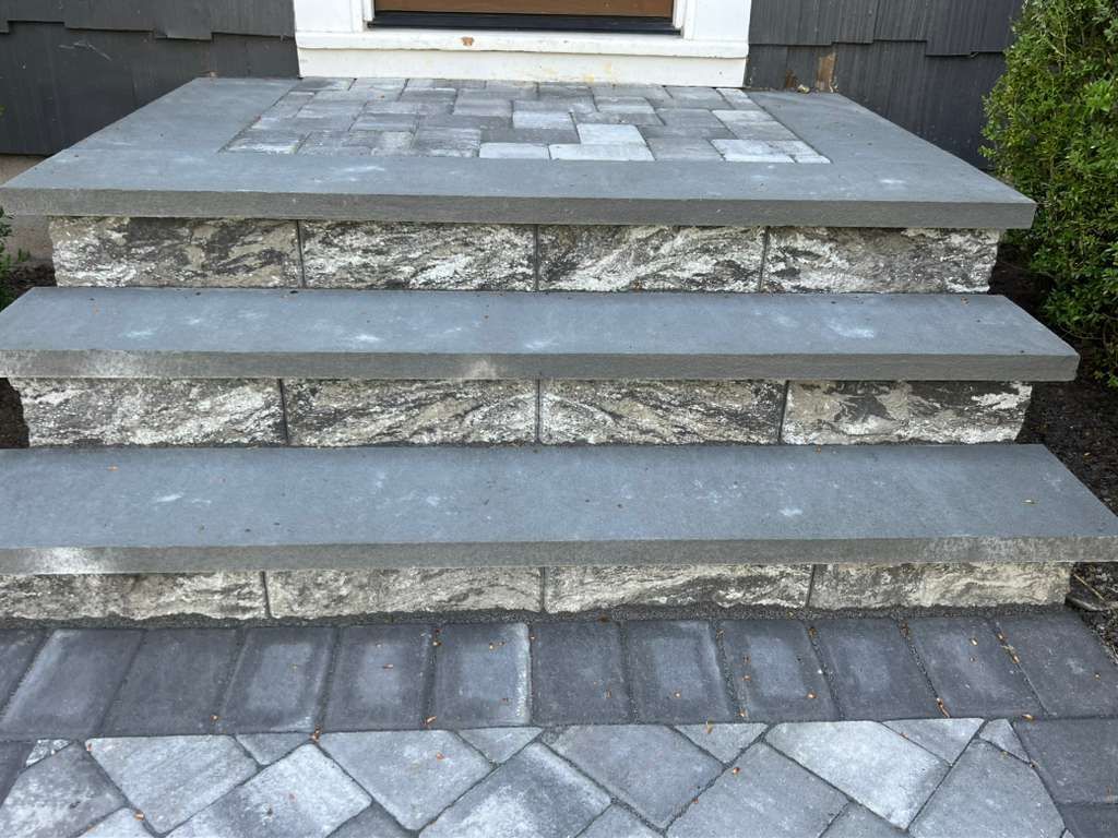 Front steps made of gray stone leading to a porch with patterned pavers and gravel sides
