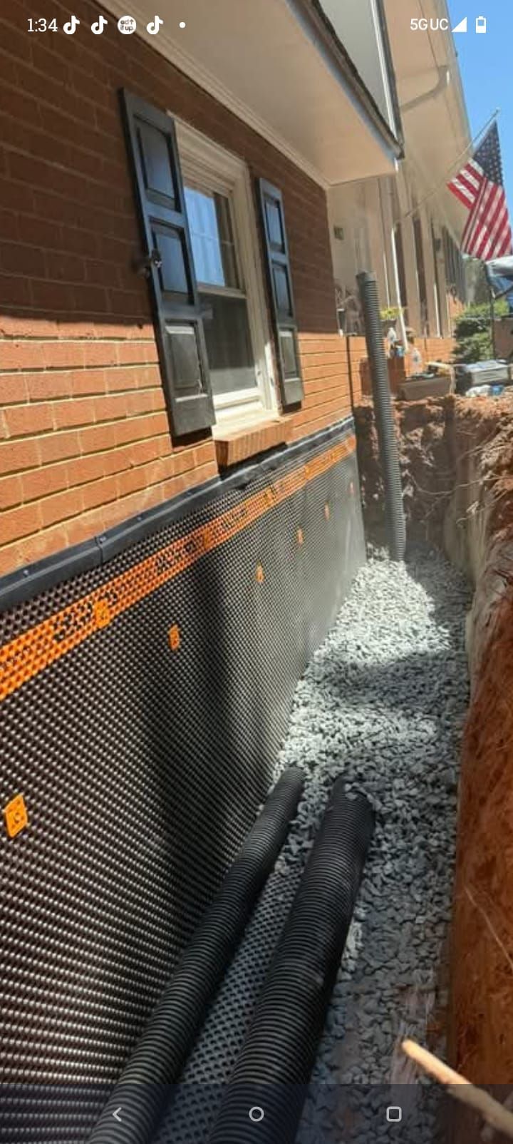 Narrow side yard beside brick house, with overflowing water spraying from ground near black drain pipes.