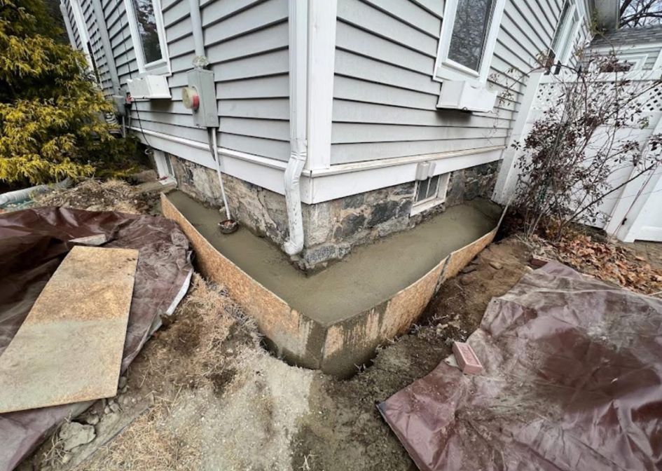 Corner of a house with a cracked concrete foundation and exposed soil near siding and basement windows.
