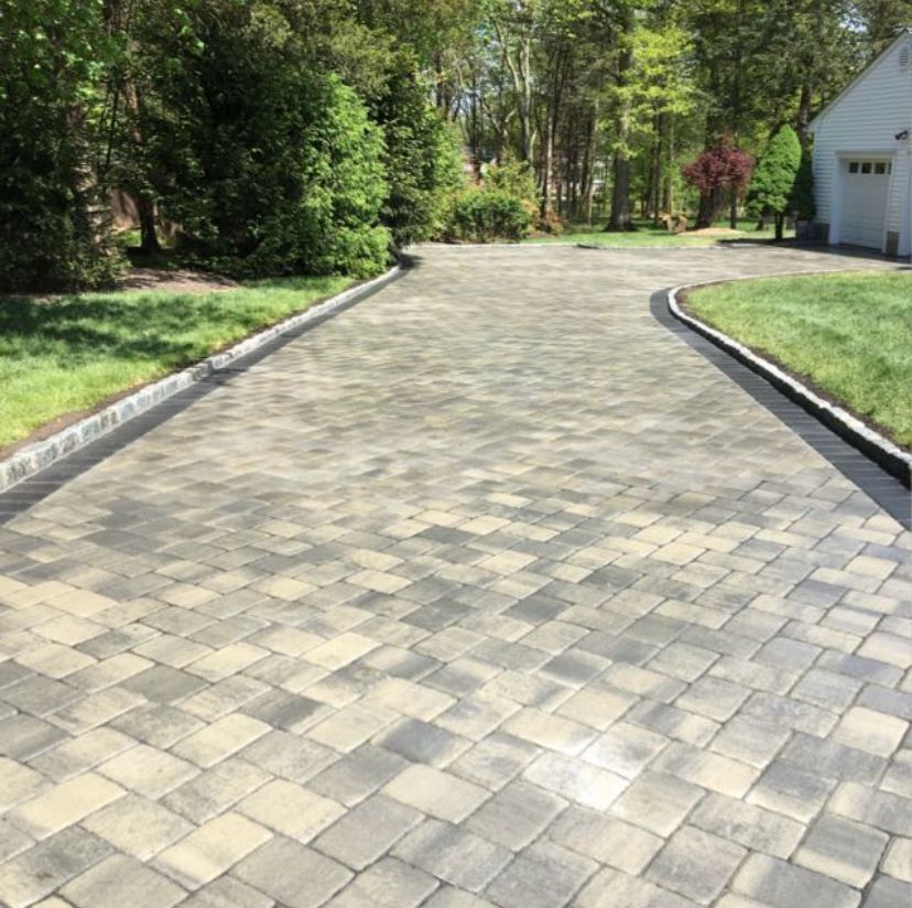 Curved gray paver driveway bordered by grass and trees, leading to a white garage