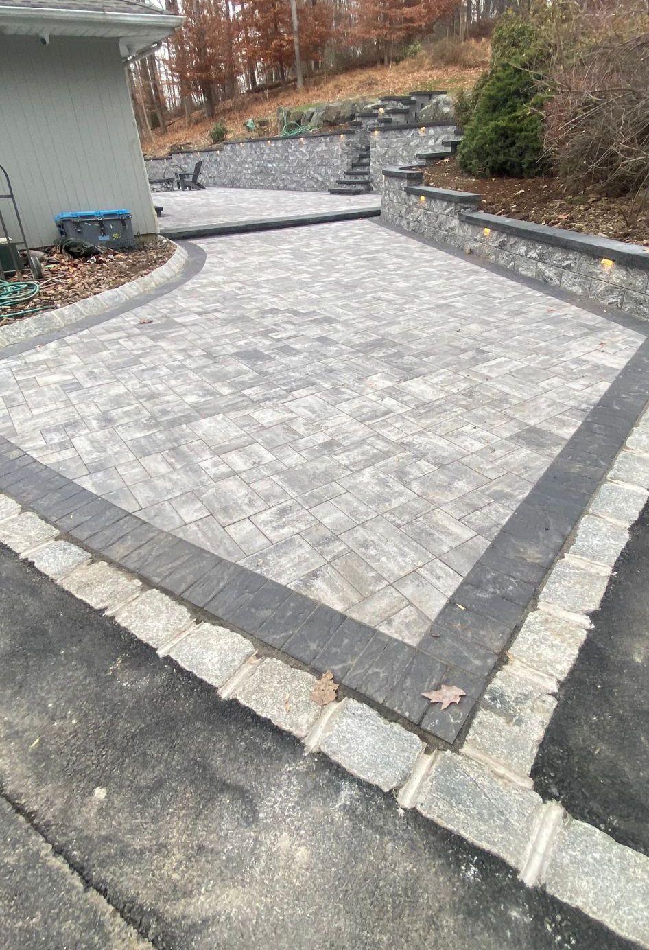 Stone-paver patio with gray border beside a house and stone retaining wall in a yard.
