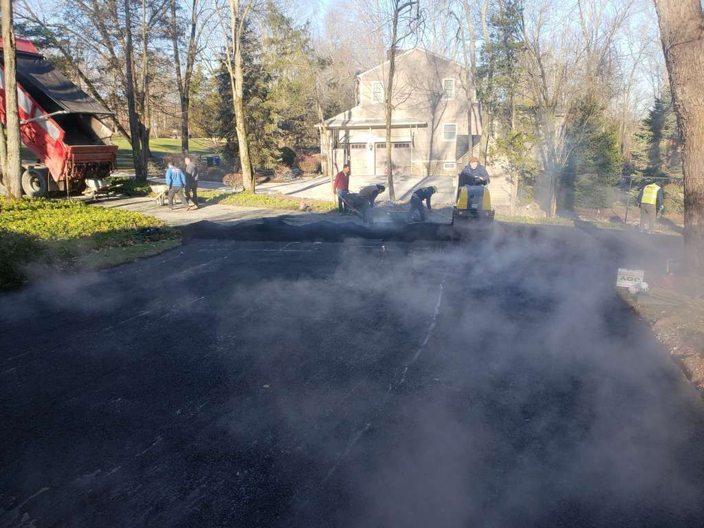 Road paving crew working on a smoky residential street with houses and bare trees nearby