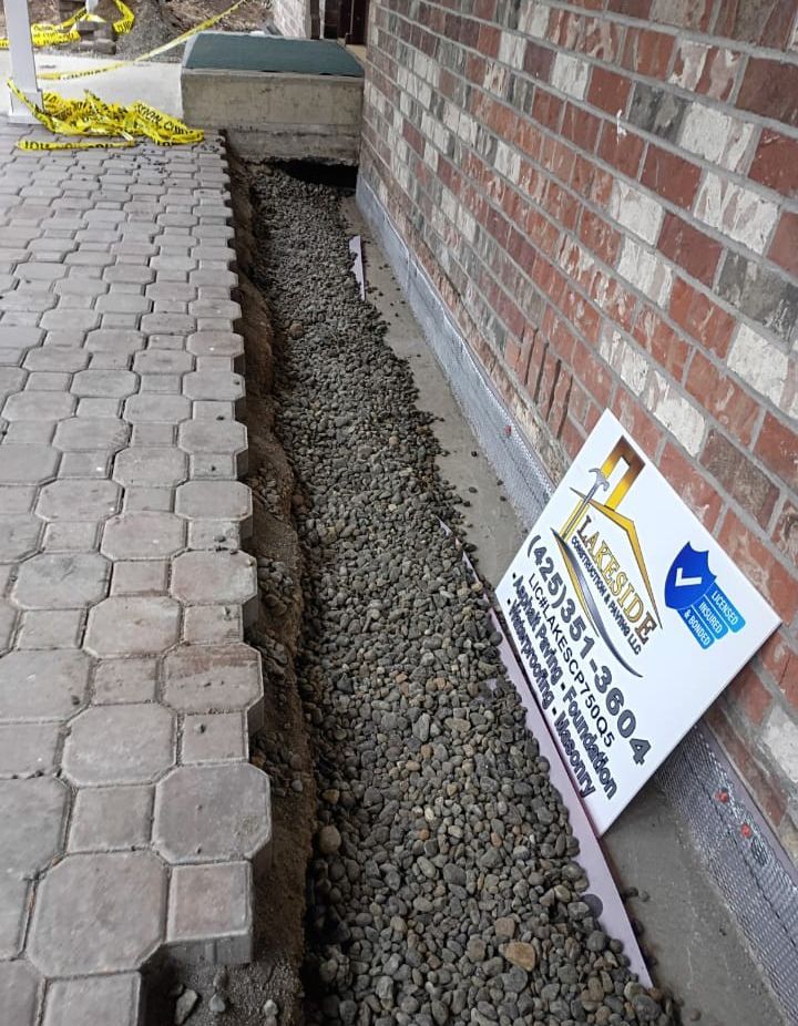 A brick wall beside a paver walkway with a narrow gravel trench and a construction sign leaning against it.