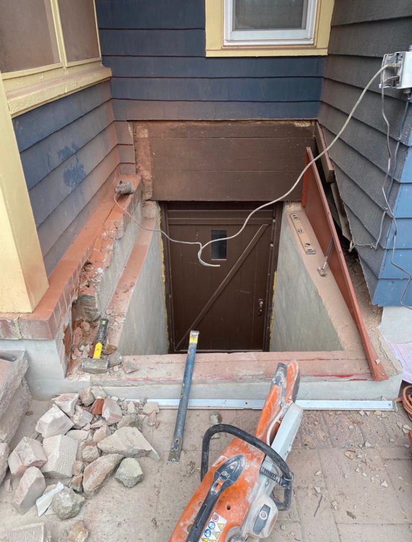 Open basement stairwell beside house exterior, with exposed dirt, bricks, and tools at the edge