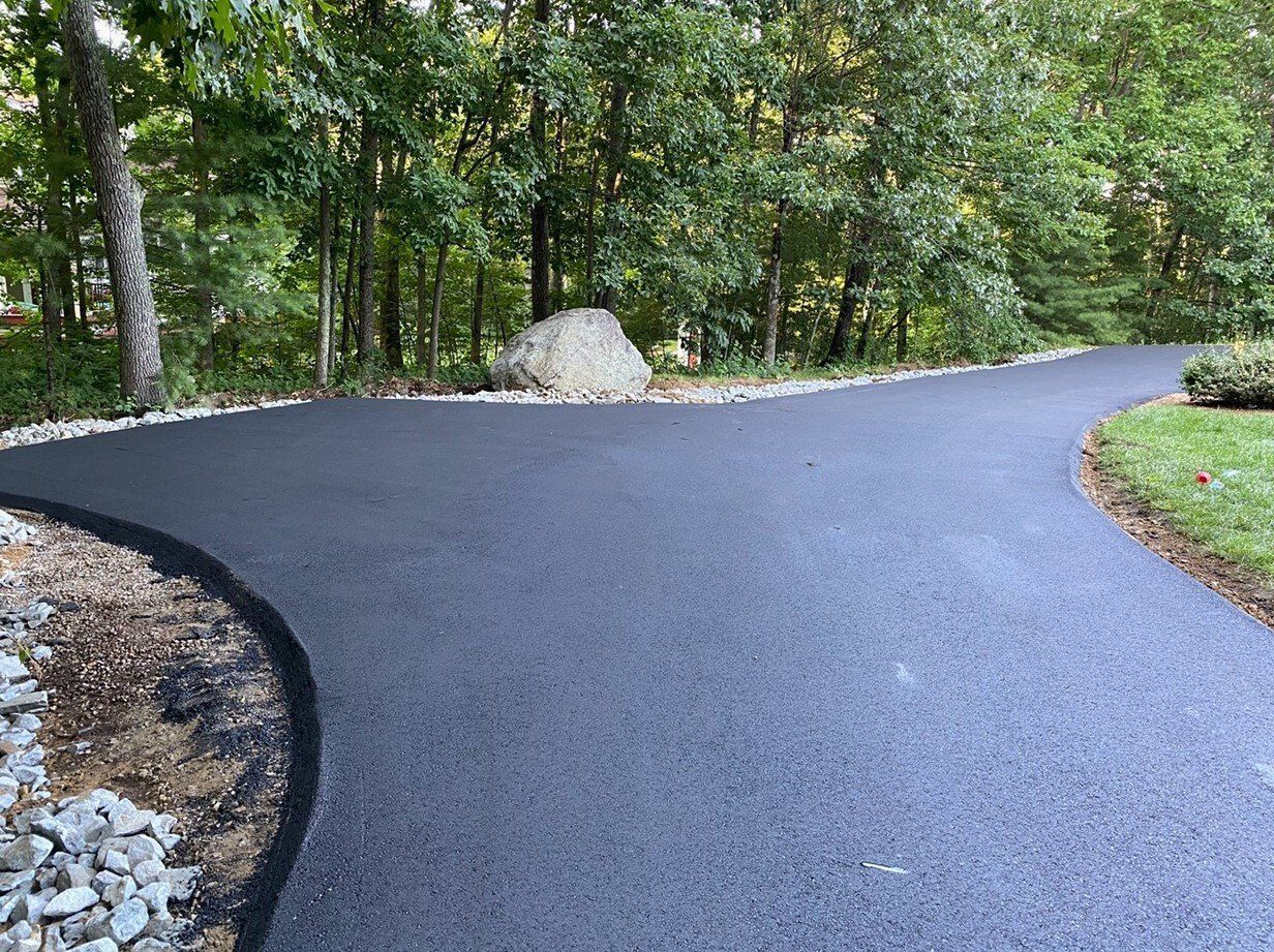 Curving dark asphalt path through a wooded area with a large rock beside it