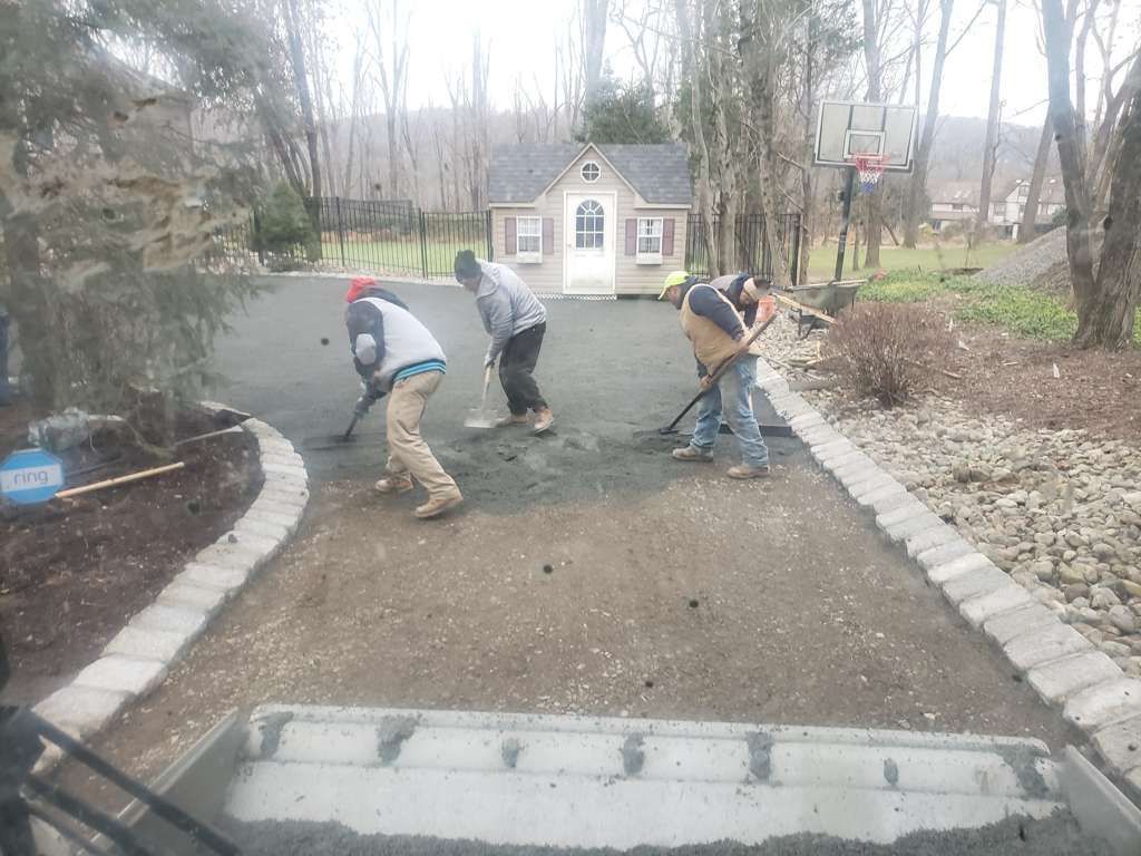 Three workers shoveling and smoothing gravel on a driveway in a yard, with a small house in the background