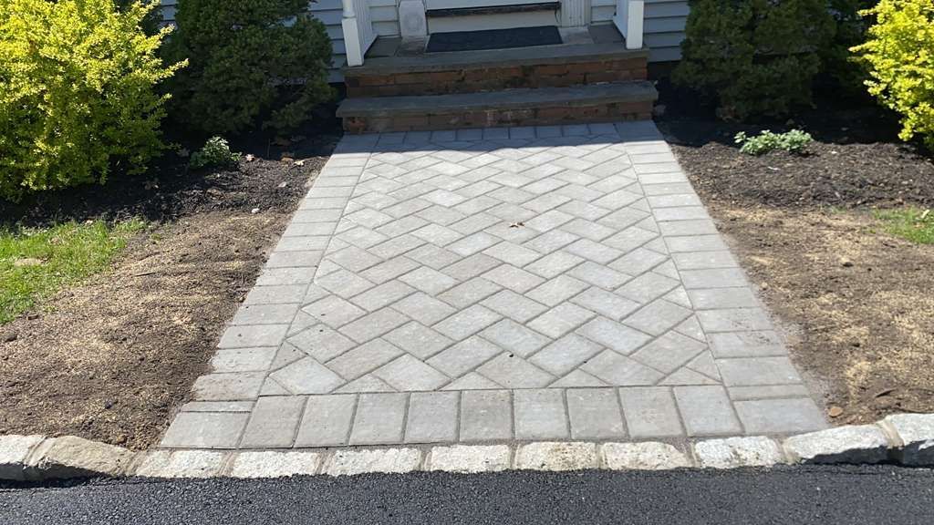 Stone paver walkway leading to a house entrance, bordered by mulch and shrubs.