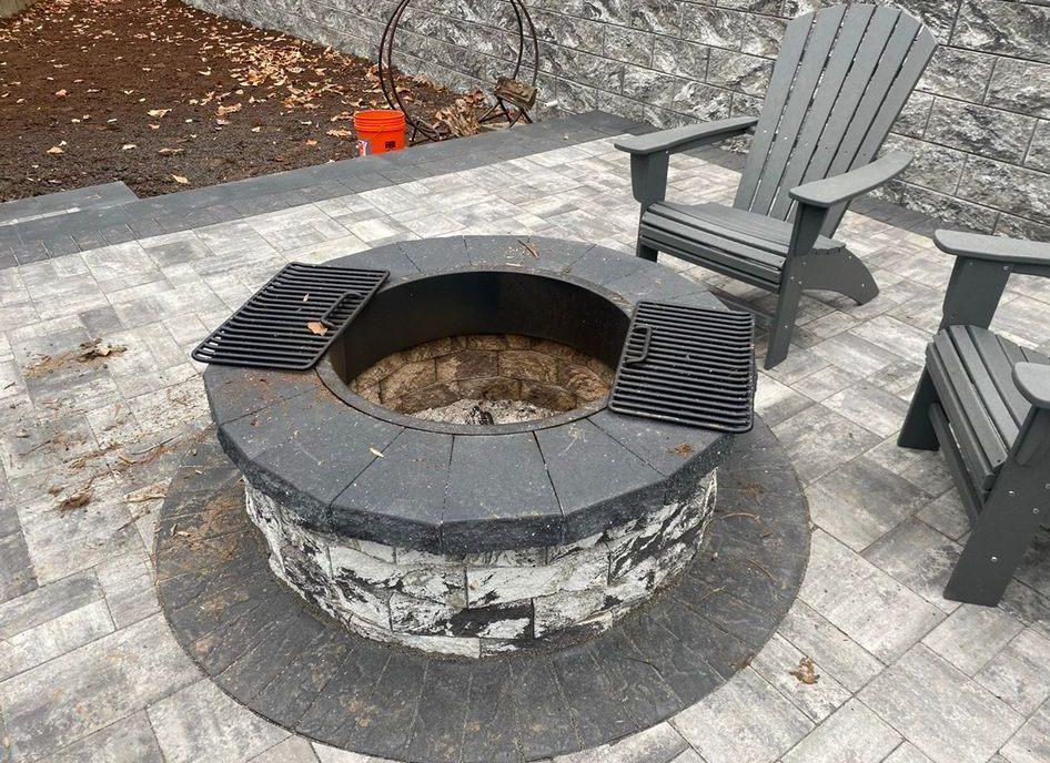 Stone fire pit with black grill grates and gray Adirondack chairs on a paved patio