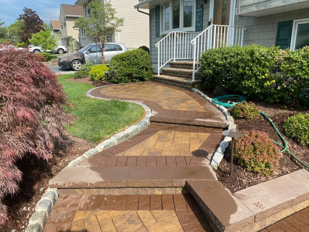 Curved brick walkway with steps leading to a white porch in a suburban front yard, bordered by shrubs and grass