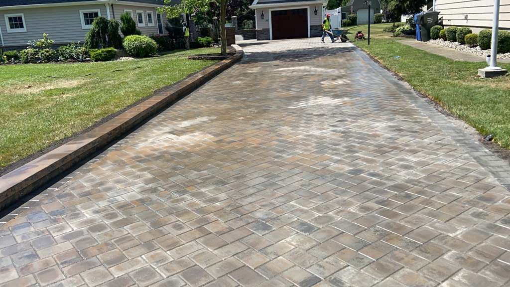 Brick driveway leading to a house, bordered by grass and black edging on both sides.