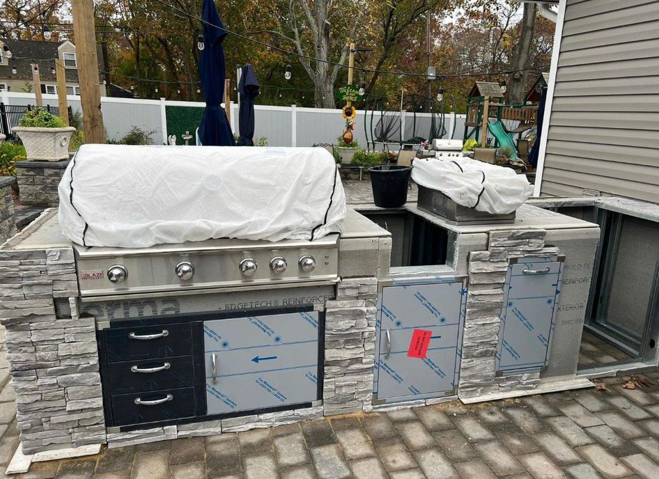 Covered outdoor stainless-steel grill and cabinets on a patio with stone pavers, next to a house.