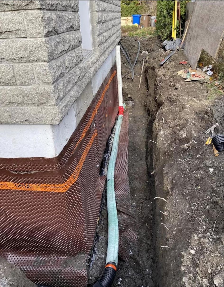 House foundation with waterproofing mesh and exposed drain pipe beside an excavated trench.